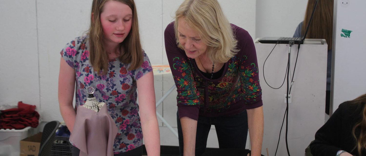 A mentor and a Teen Squad member review a sketch of a dress in front of a miniature dress form with a dress on it