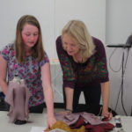 A mentor and a Teen Squad member review a sketch of a dress in front of a miniature dress form with a dress on it