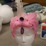 A pink turban style headdress with a white feather and purple jewels