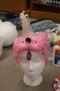 A pink turban style headdress with a white feather and purple jewels